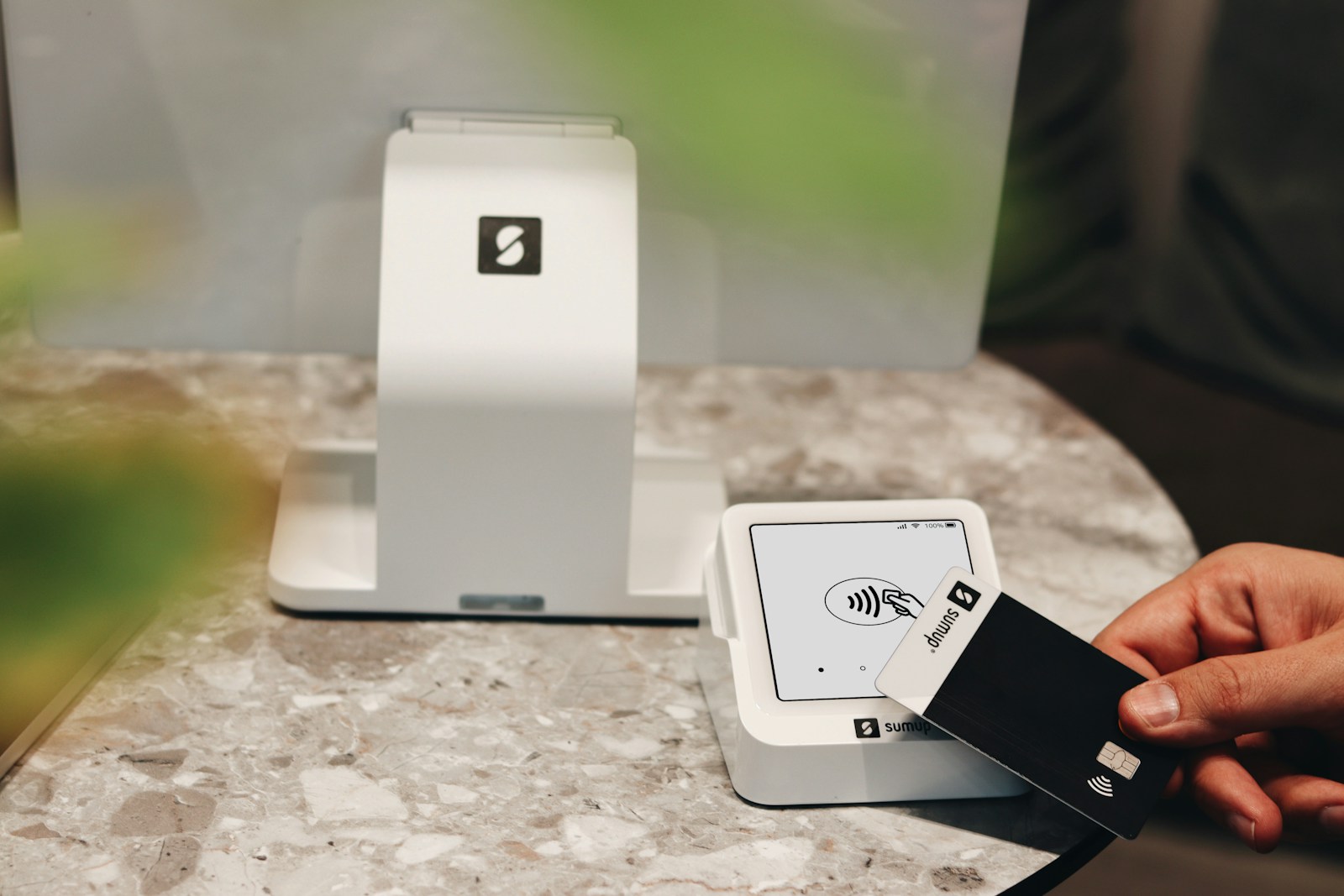 A SumUp Solo card reader is being used to make a contactless payment with a SumUp card. The device is placed on a sleek, modern marble counter, enhancing the overall minimalist and professional aesthetic. The SumUp POS system behind the card reader emphasizes the streamlined and easy-to-use technology, ideal for cafes, boutiques, and other small businesses that value both function and design. This setup reflects a commitment to modern payment solutions with a stylish touch.